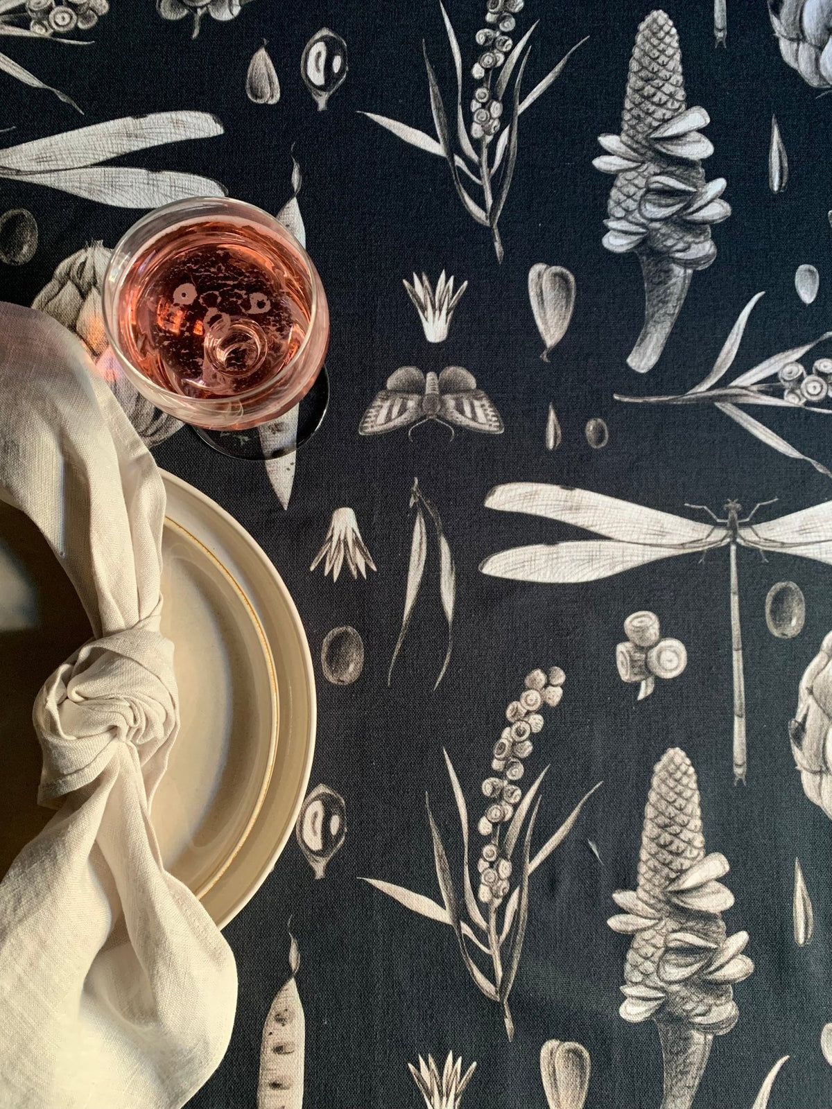 3. Close-up of Seeds Carbon tablecloth pattern with dragonfly and corn motifs, accompanied by a glass of rosé and a knotted napkin