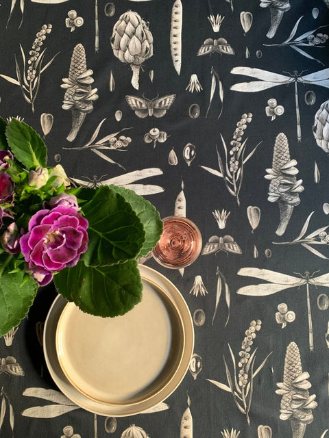 2. Top view of Seeds Carbon print tablecloth with floral and insect motifs, featuring a plate and pink flowers