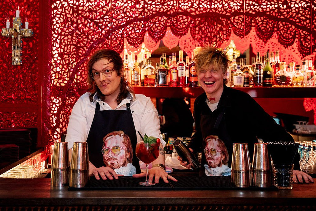 1. Two men behind a bar wearing Katrin Valgemäe's 'Pervert in the Kitchen' aprons, surrounded by colorful decor