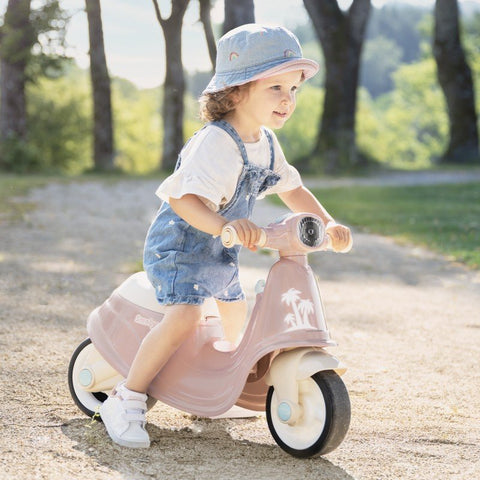 1. Child wearing denim overalls and hat riding pink Smoby retro ride-on scooter in park setting