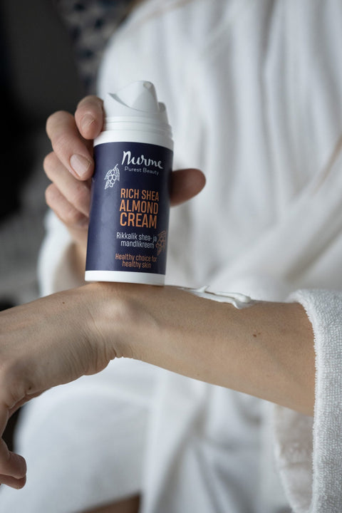 1. Person applying Nurme Rich Shea Almond Cream to arm, holding 100ml bottle with dark blue label, in a cozy indoor setting