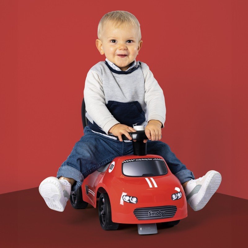 1. Toddler boy sitting on Smoby red ride-on car with horn, wearing casual outfit, against red background