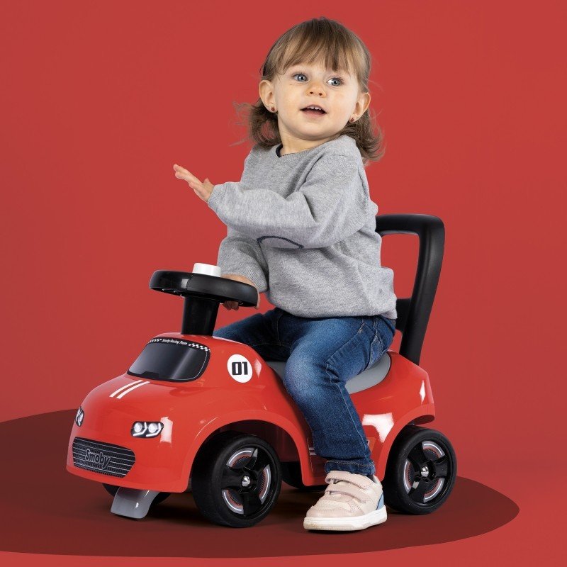 4. Toddler girl on Smoby red ride-on car with horn, wearing grey sweater and jeans, against red background