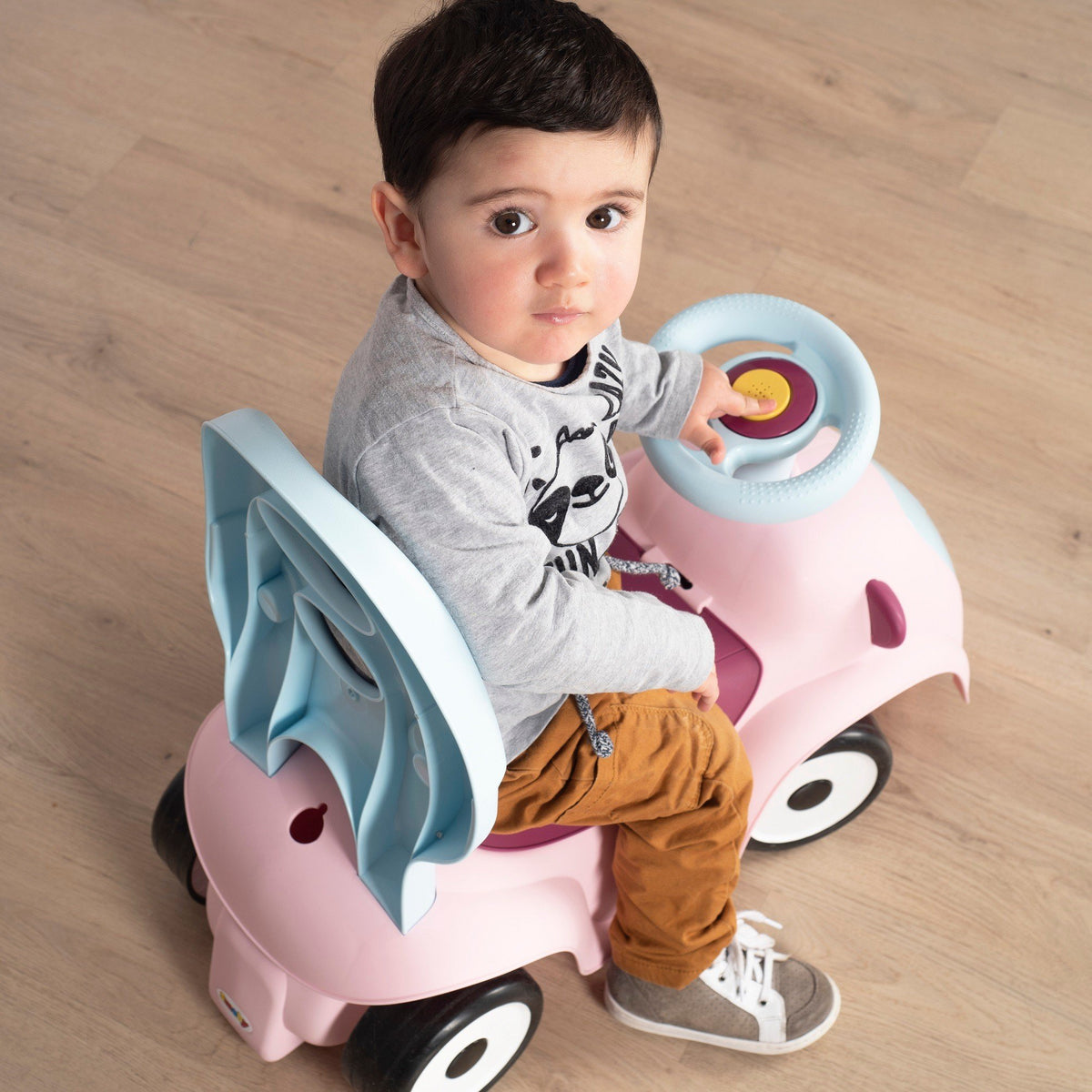 11. Child sitting on Smoby Ride-on Maestro 3in1 in pink, holding steering wheel, wearing a grey shirt