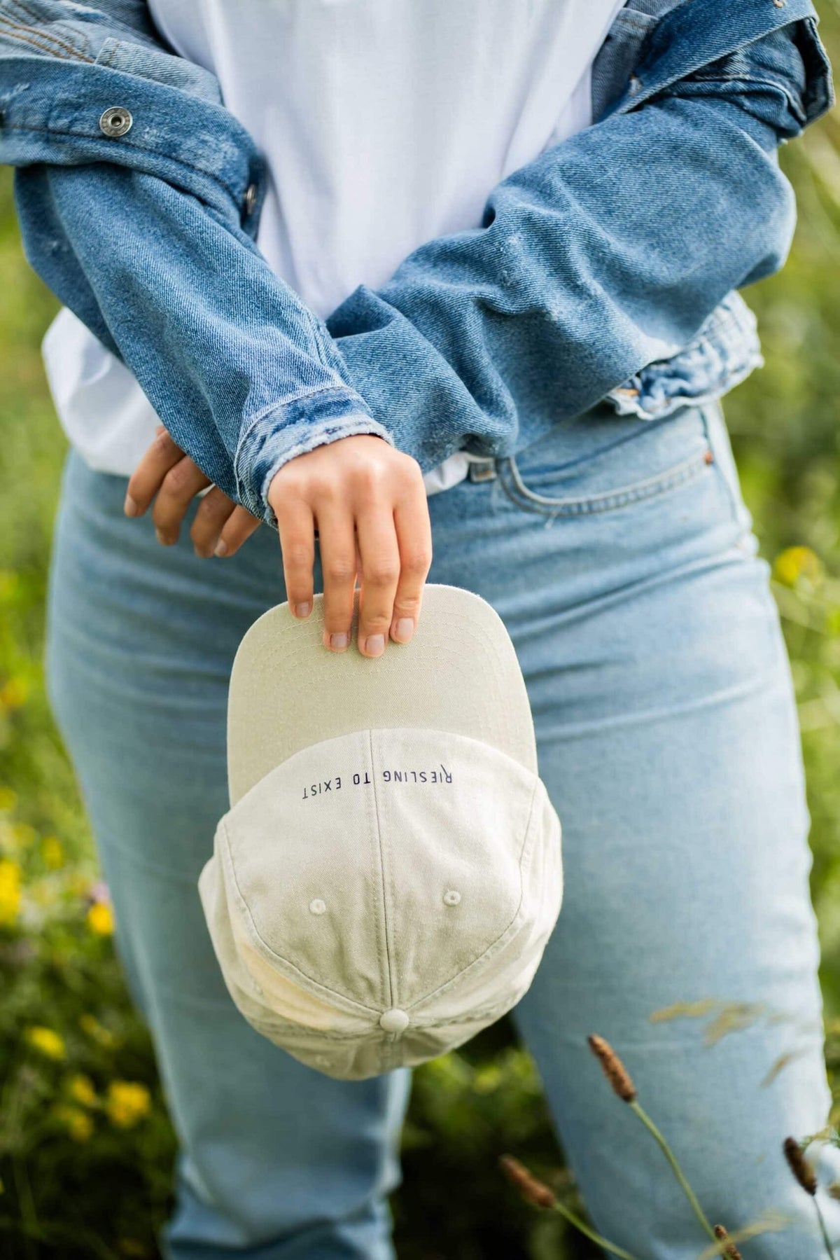 1. Woman holding beige 'Riesling to Exist' cap in outdoor setting wearing denim jacket and jeans