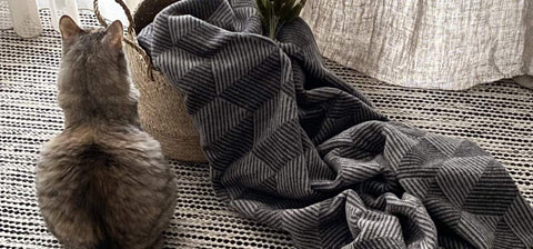 1. Gray cat sitting next to Labbvenn Rino Blanket in grayish tones with geometric patterns, draped over a woven basket on a textured rug in a cozy interior setting