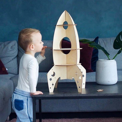 1. Child standing next to Cut It Now rocket-shaped wooden shelf with star cut-outs in a living room setting