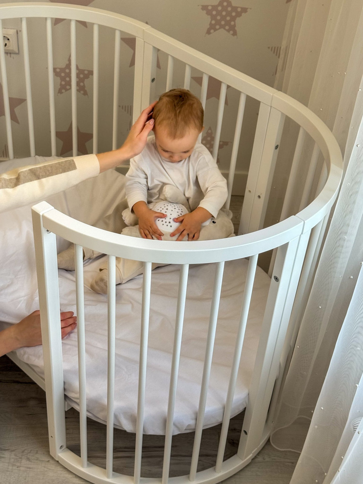 14. Baby sitting in white round crib with a woman’s hand reaching in