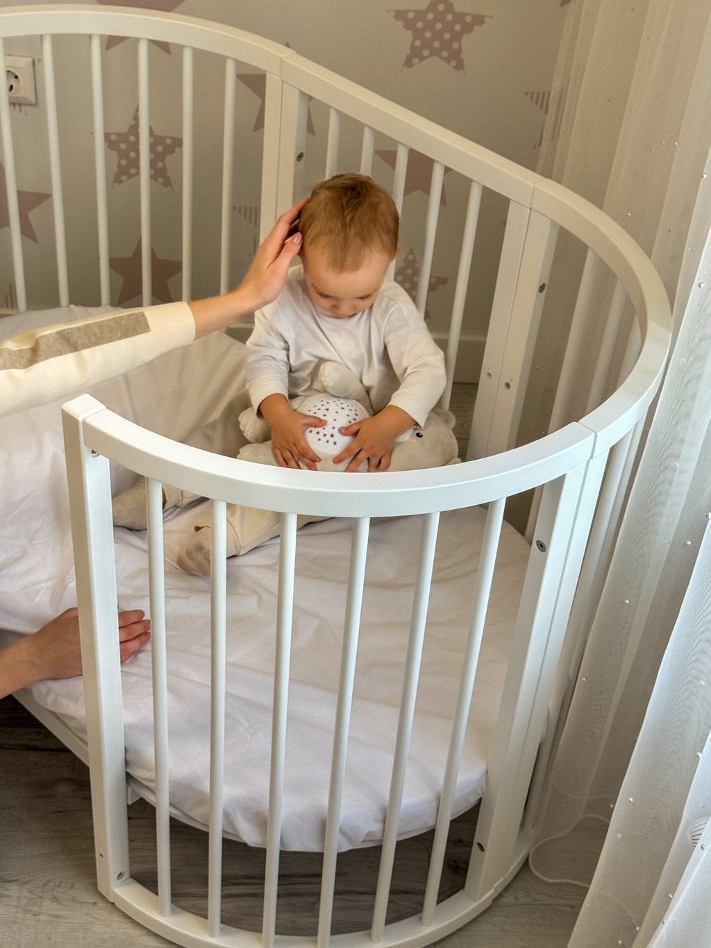 5. Baby sitting in white round crib with hand reaching in, highlighting child-safe design