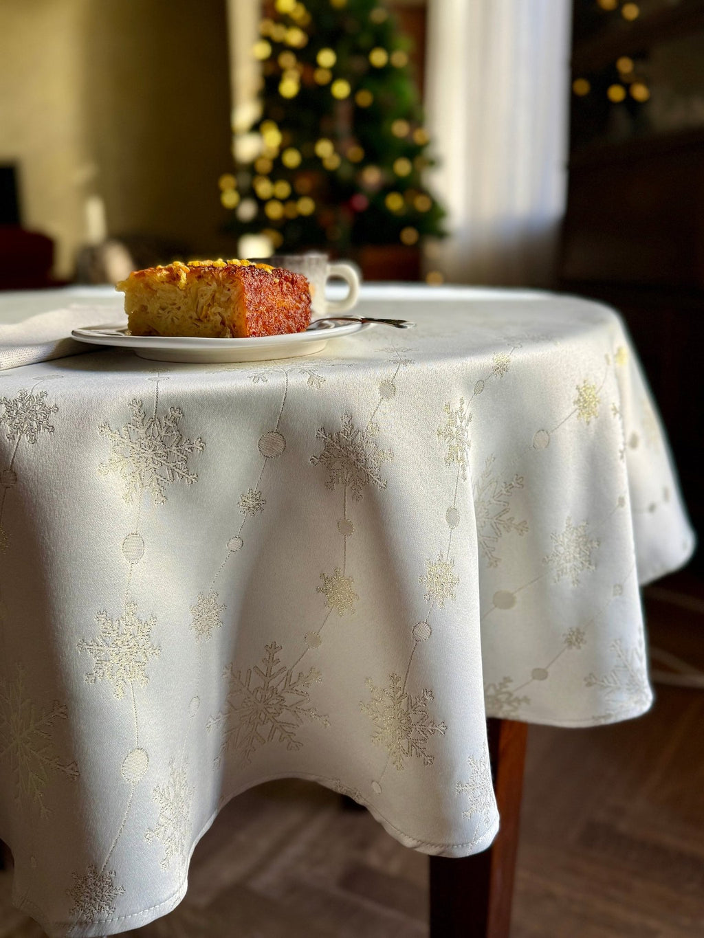 1. Festive dining room setting with round table featuring Hortensias Home Golden Snowfall tablecloth, subtle golden snowflake design, slice of cake on plate, and Christmas tree in background