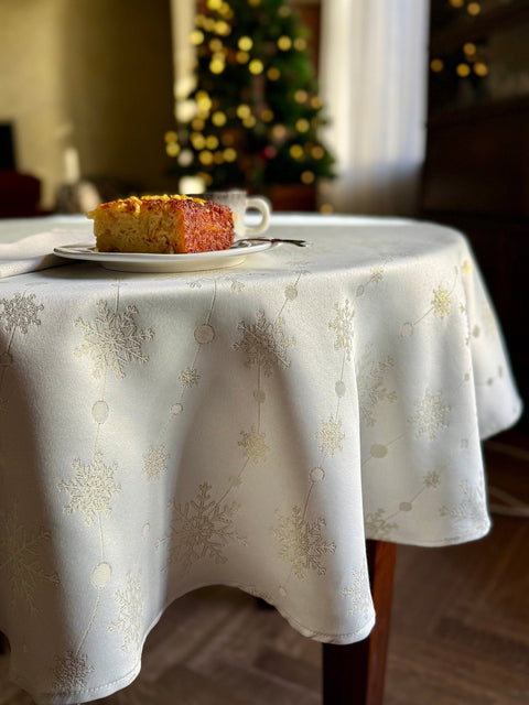 1. Festive dining room setting with round table featuring Hortensias Home Golden Snowfall tablecloth, subtle golden snowflake design, slice of cake on plate, and Christmas tree in background
