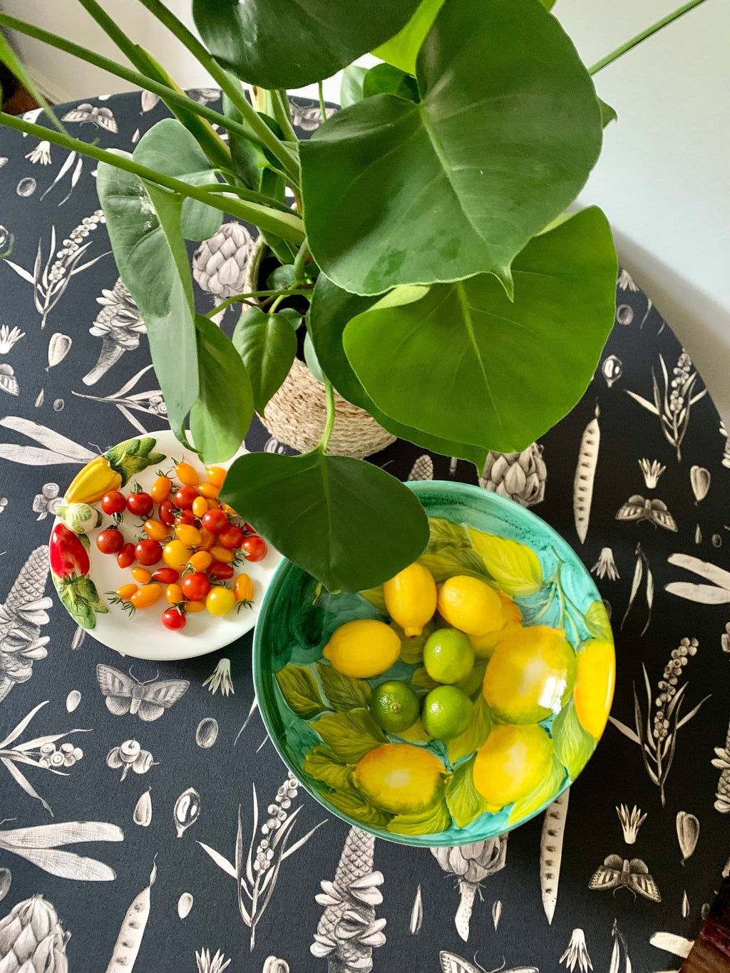 6. Round table with black Seeds Carbon print tablecloth, featuring artichoke and pea pod designs, topped with a bowl of lemons and limes, and a green plant