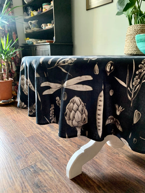 5. Black Seeds Carbon print tablecloth on a round table, showcasing artichoke and dragonfly designs, in a dining room with wooden floor and potted plants