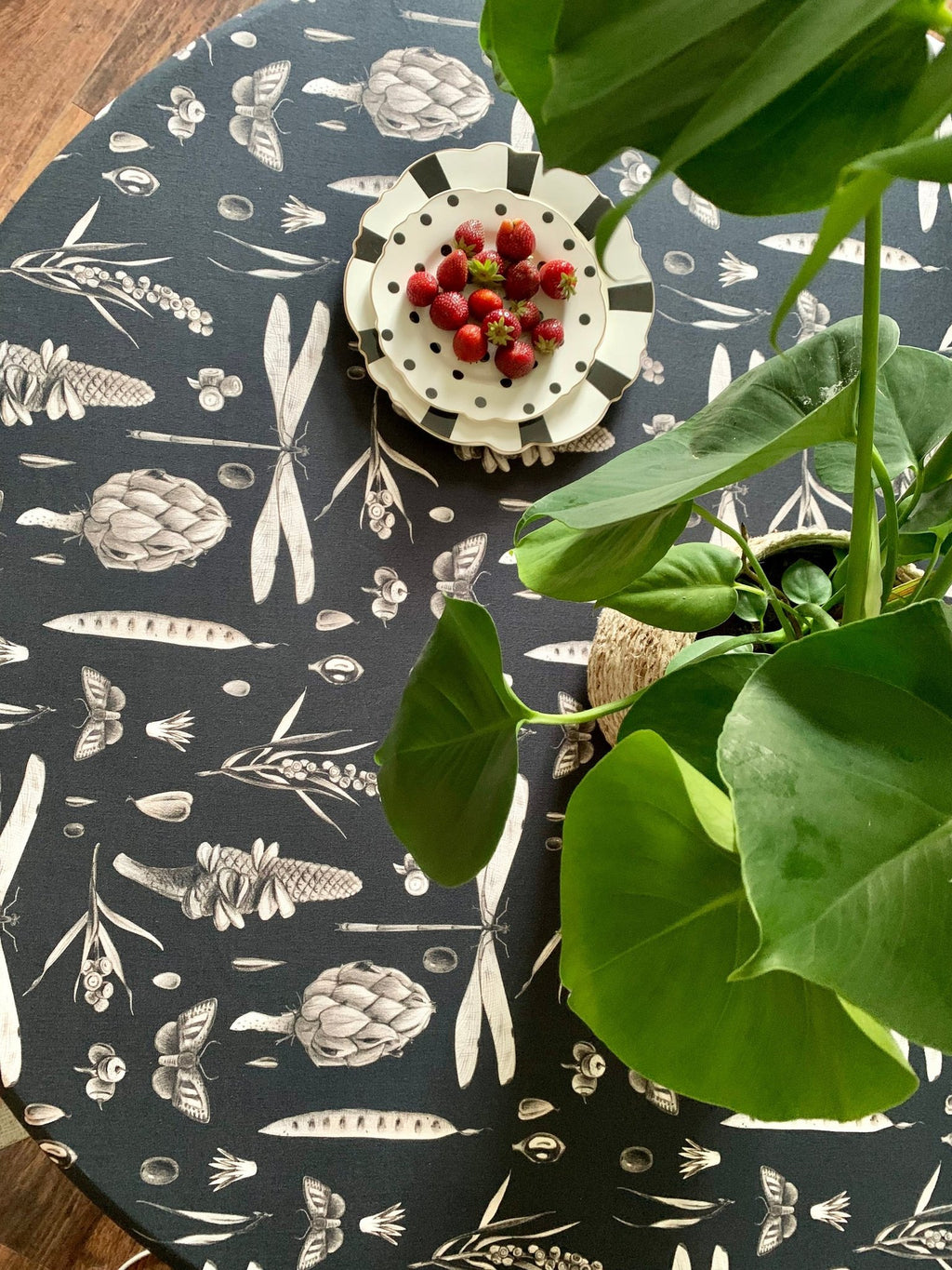 1. Round table with black Seeds Carbon print tablecloth, featuring artichoke and pea pod designs, topped with a plate of strawberries and a green plant in a dining room setting
