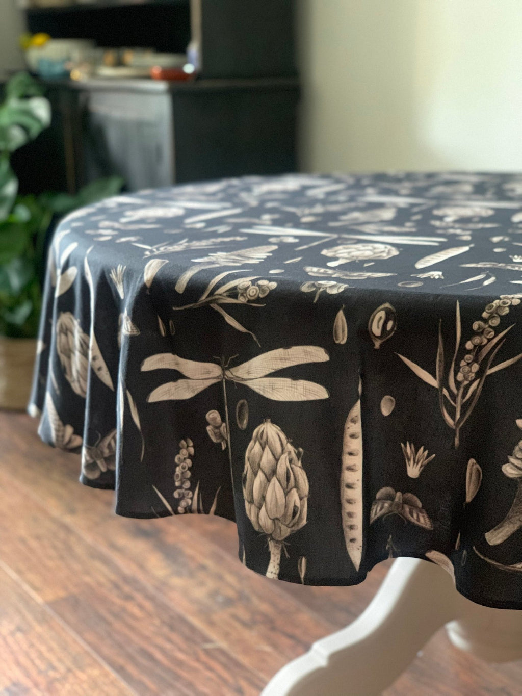 3. Black Seeds Carbon print tablecloth draped over a round table, highlighting artichoke and dragonfly designs in a dining room with wooden floor