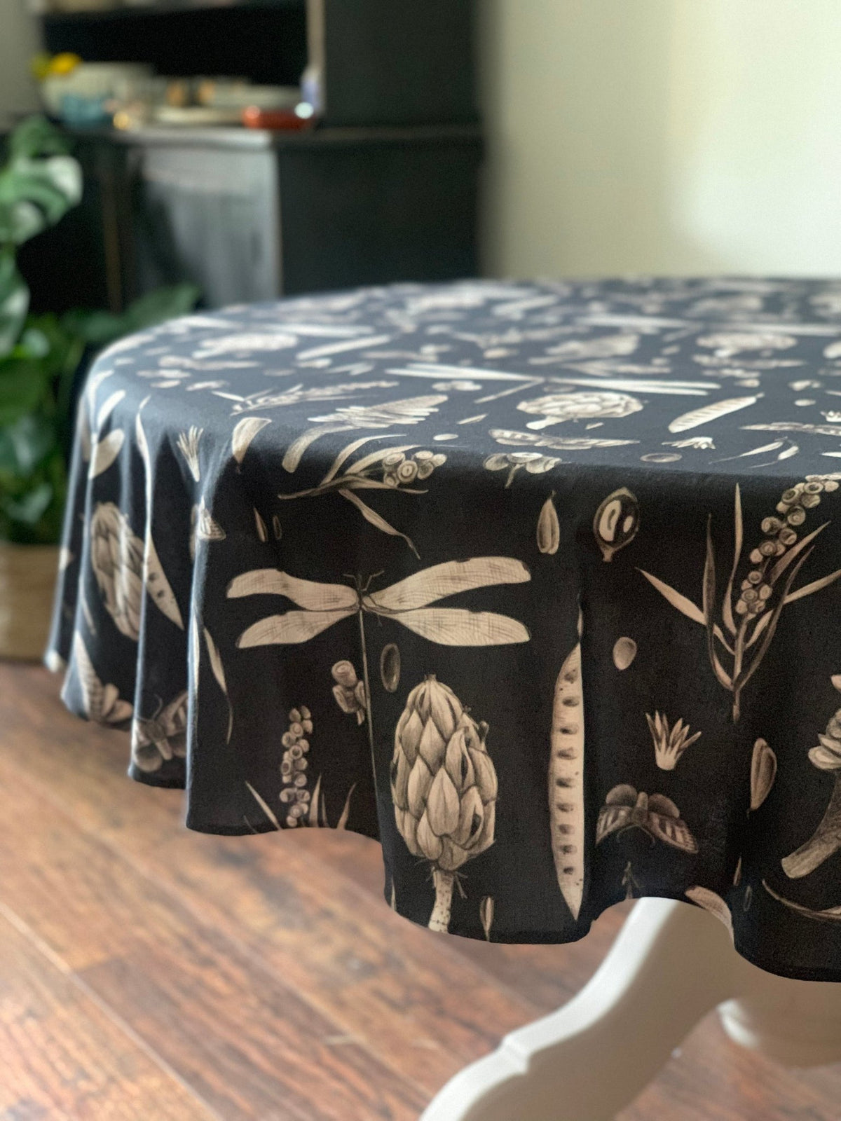 3. Black Seeds Carbon print tablecloth draped over a round table, highlighting artichoke and dragonfly designs in a dining room with wooden floor