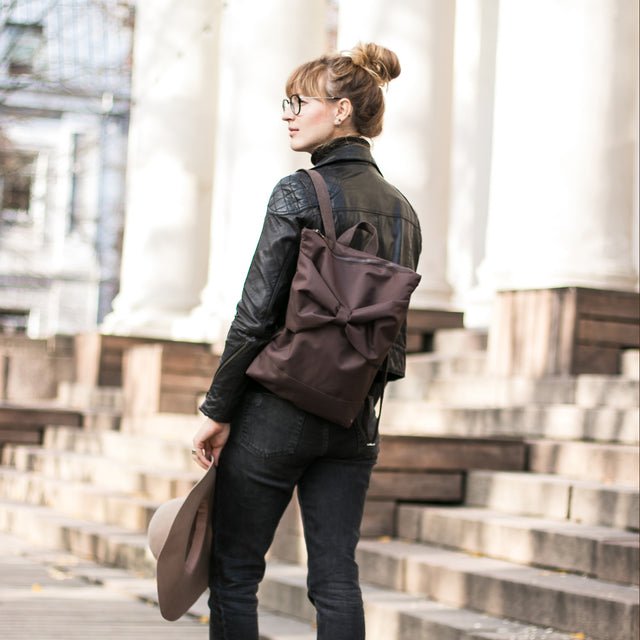 2. Woman showcasing MUNI brown backpack with bow, worn on back in city environment