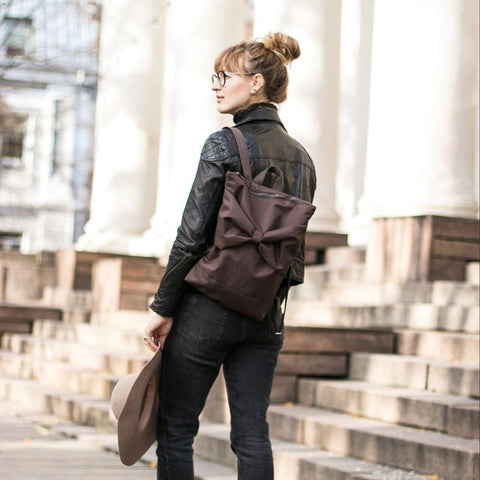 2. Woman showcasing MUNI brown backpack with bow, worn on back in city environment