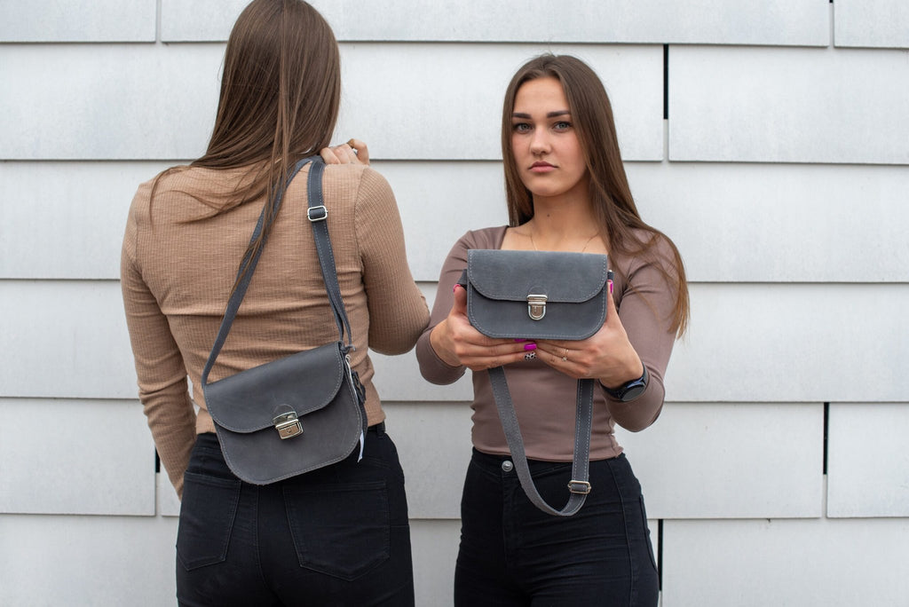 1. Two women showcasing the Zelma Kraft grey leather saddle bag, one wearing it crossbody and the other holding it, against a modern backdrop