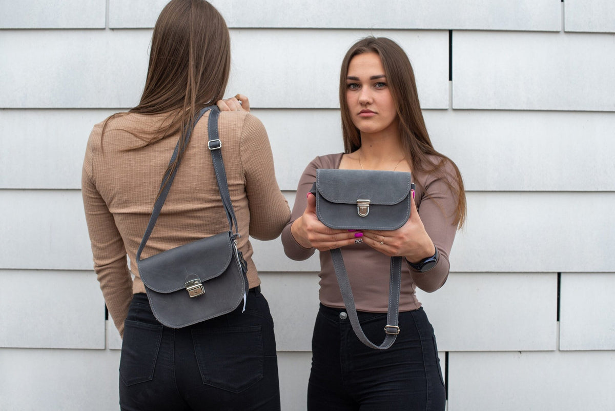 1. Two women showcasing the Zelma Kraft grey leather saddle bag, one wearing it crossbody and the other holding it, against a modern backdrop