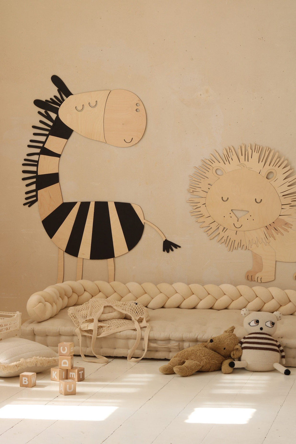 2. Wooden zebra and lion wall decorations in a nursery with beige walls, surrounded by plush toys and a braided cushion