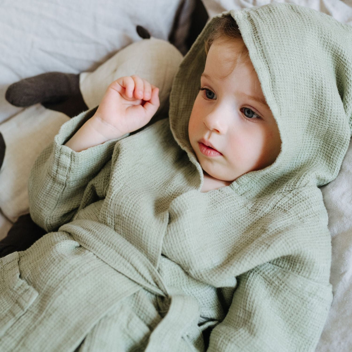 1. Child lying on bed wearing sage green linen bathrobe with hood
