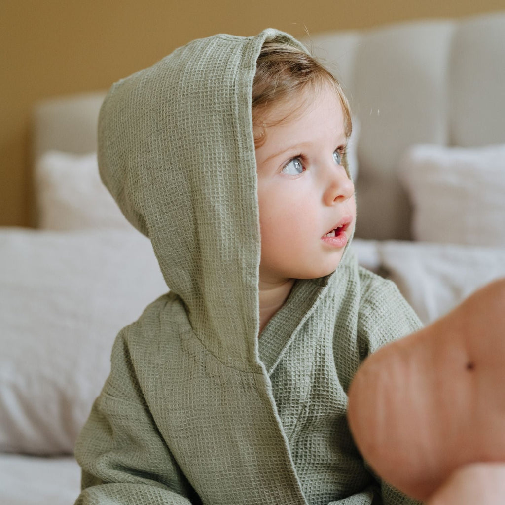 1. Child wearing sage green linen bathrobe with hood sitting on bed