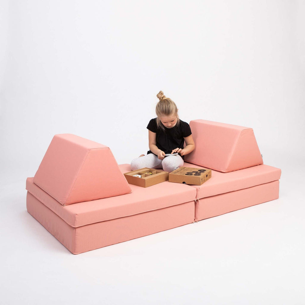 3. Girl sitting on Monboxy salmon pink play mattress set with trapezoidal pillows, playing with toys in a studio setting