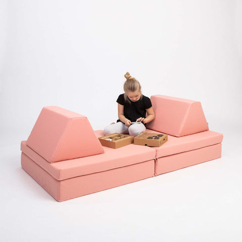 3. Girl sitting on Monboxy salmon pink play mattress set with trapezoidal pillows, playing with toys in a studio setting
