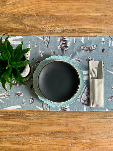 1. Table setting with Seeds Aqua table runner featuring botanical print, black plate, and green plant on wooden table