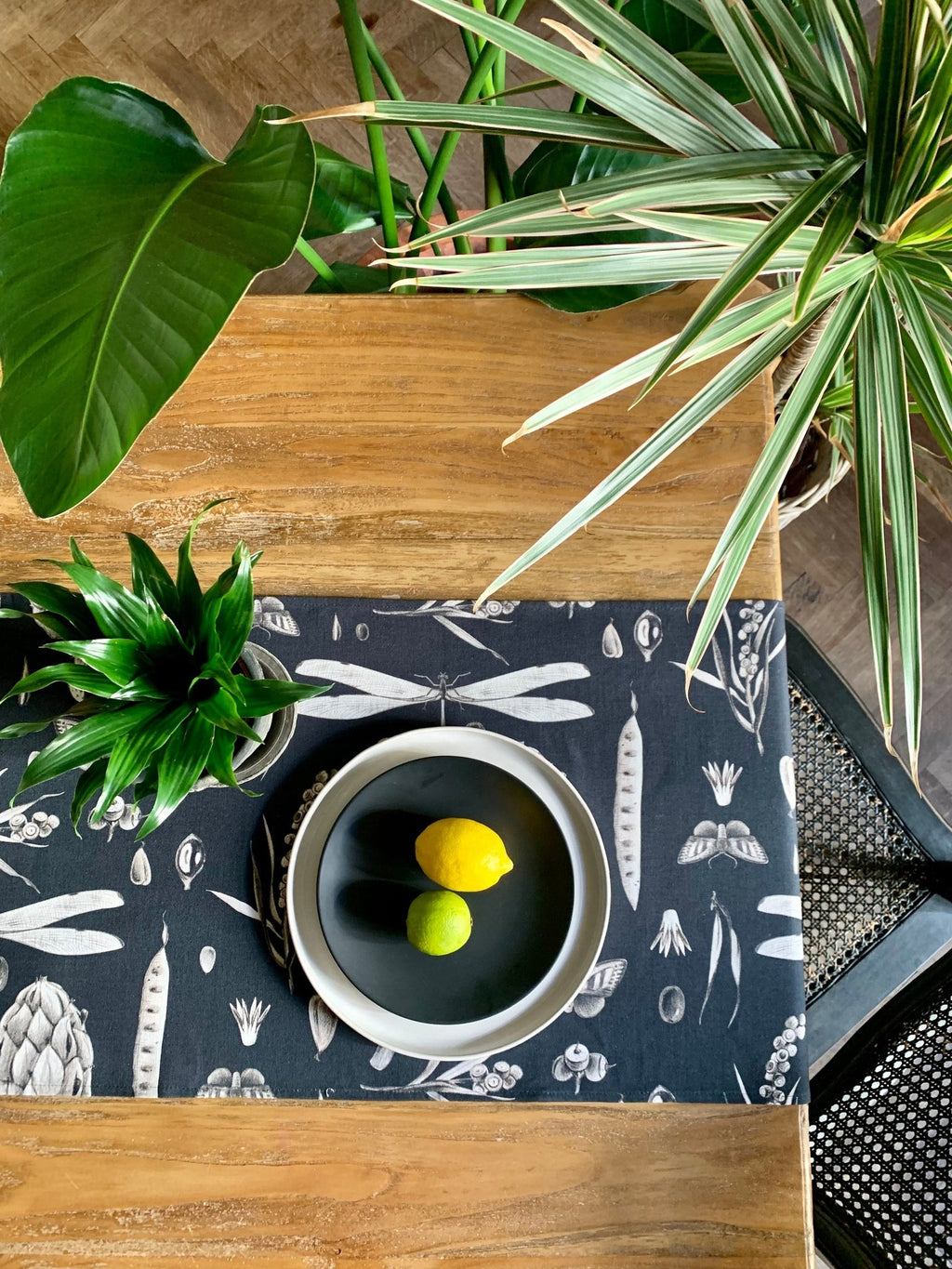 1. Dining table with Seeds Carbon print table runner in black and white, featuring a bowl with lemon and lime, surrounded by green plants