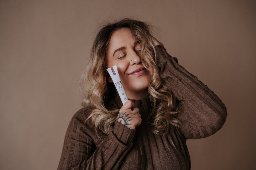 4. Woman with curly hair holding LUUV Self-Tanning Face Serum, smiling with eyes closed
