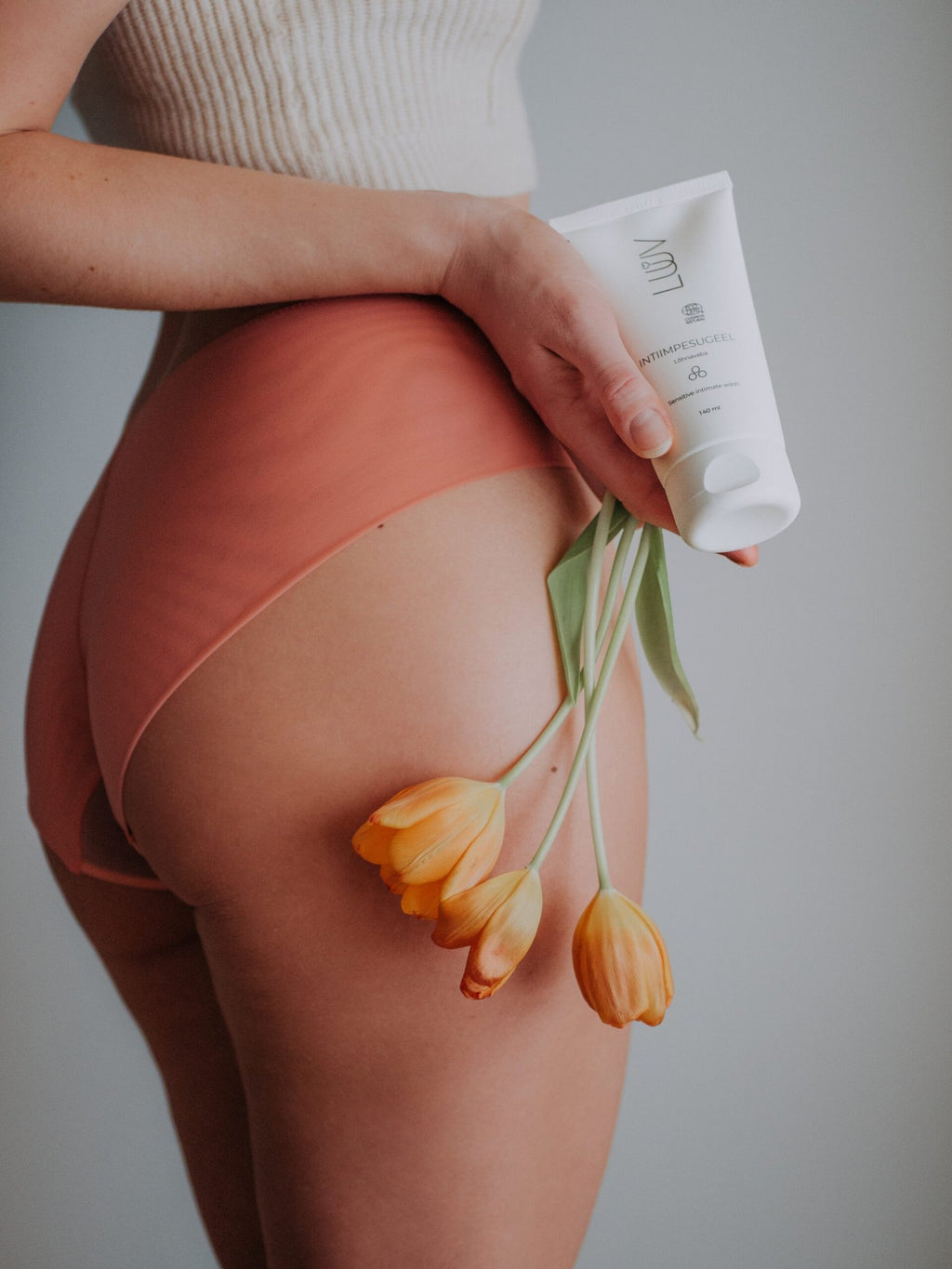 1. Woman holding LUUV Sensitive Intimate Wash and orange tulips against a neutral background, wearing peach underwear and a white top