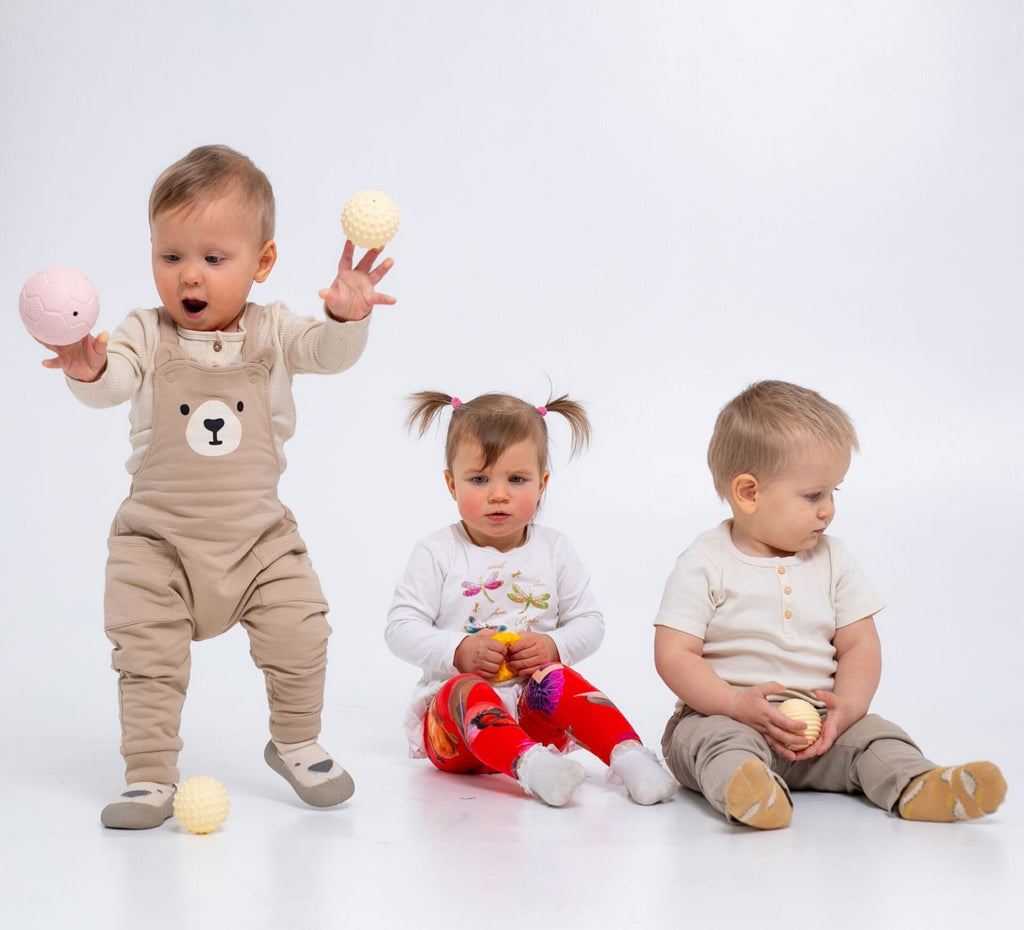 1. Three babies sitting on a white background, each holding pastel-colored sensory balls, wearing various outfits, encouraging group play and motor skills.