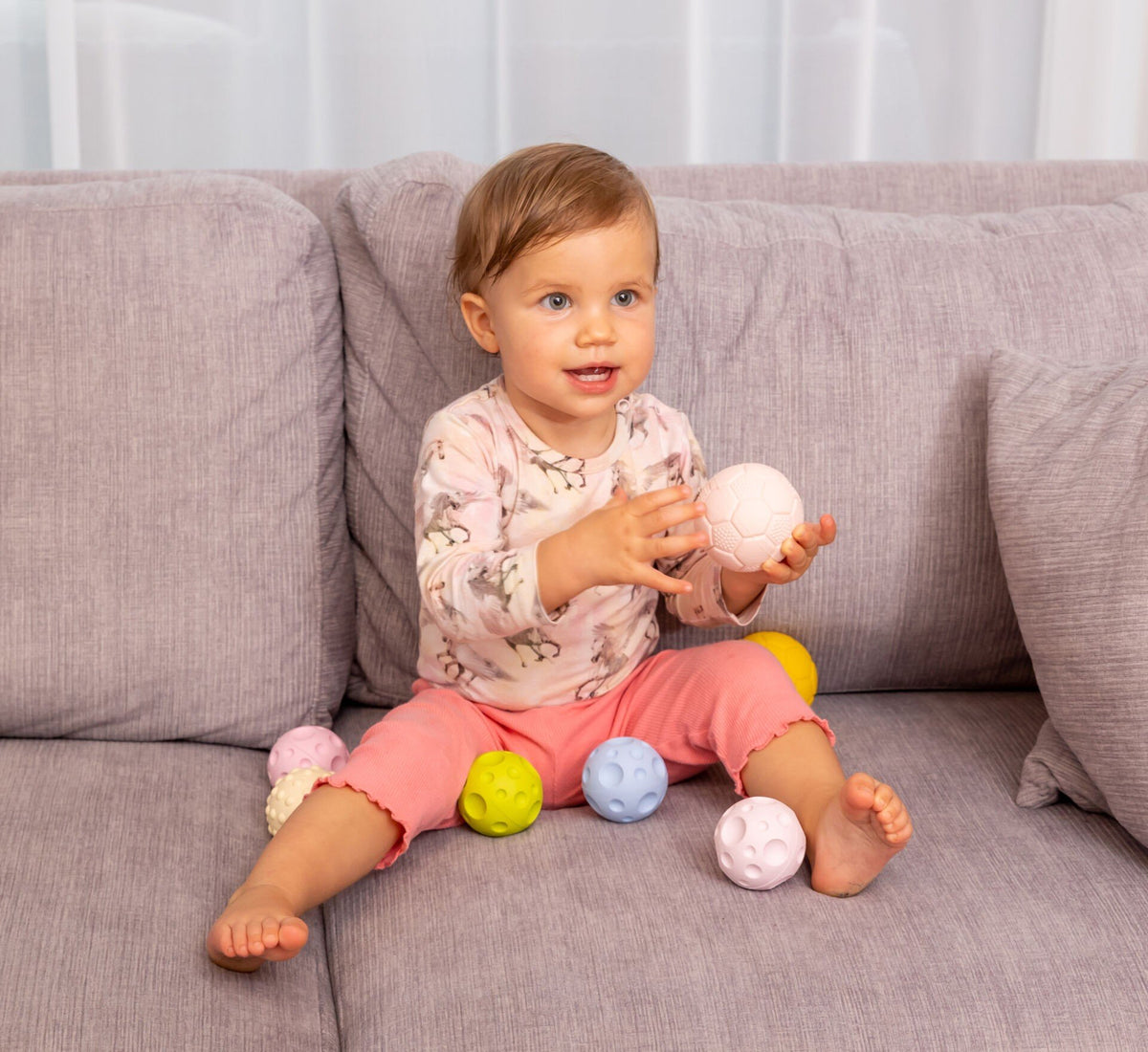 1. Baby sitting on a grey couch playing with pastel-colored sensory balls, wearing a pink outfit, promoting early motor skills development.