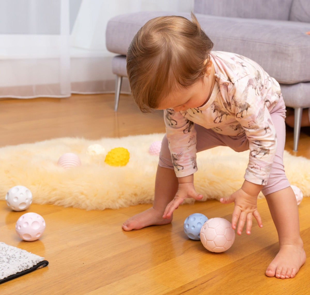 1. Baby in a pink outfit bending over to pick up pastel-colored sensory balls on a wooden floor, encouraging active play and coordination.