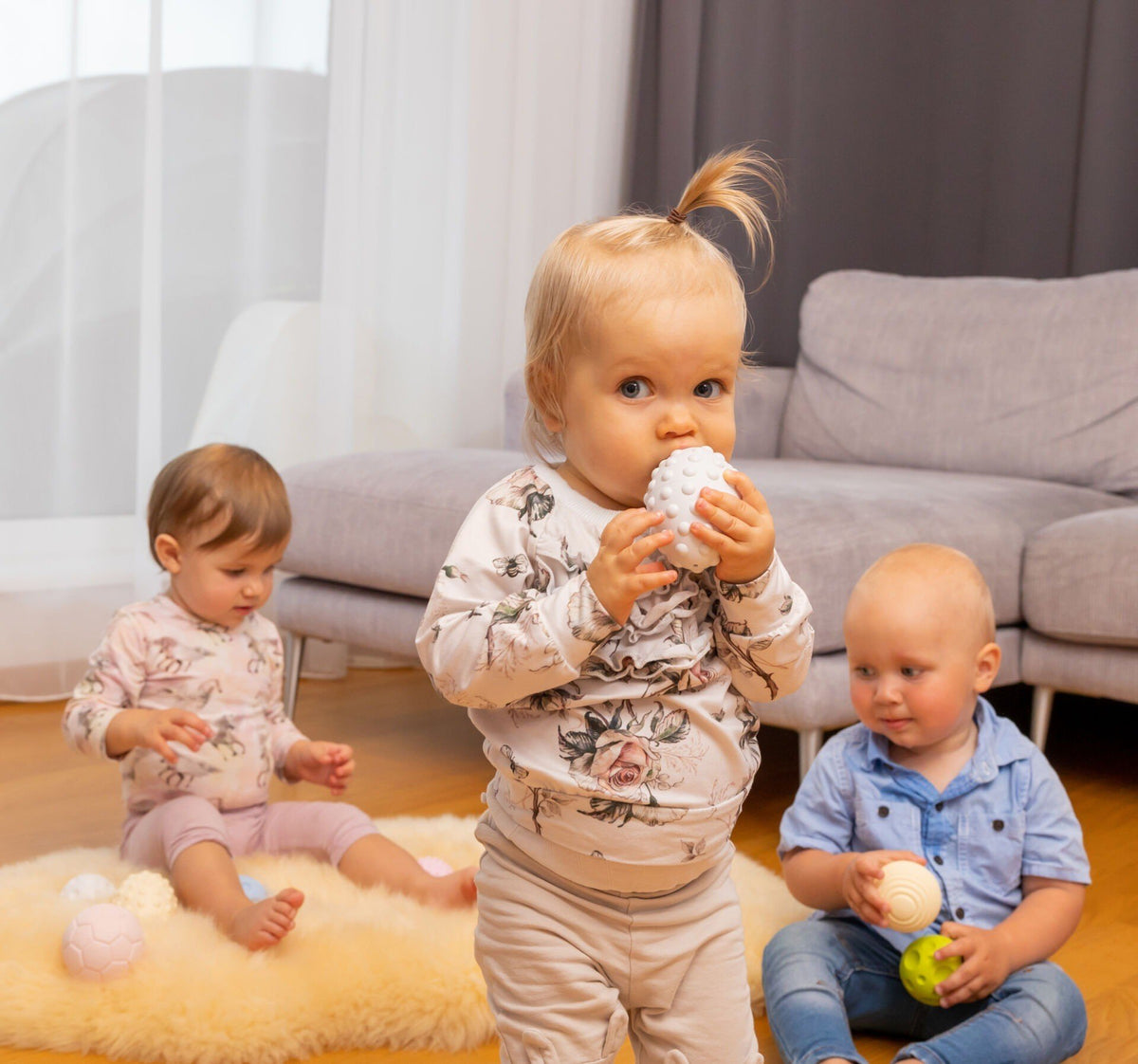 1. Three babies playing with pastel-colored sensory balls in a living room setting, promoting social interaction and sensory development.