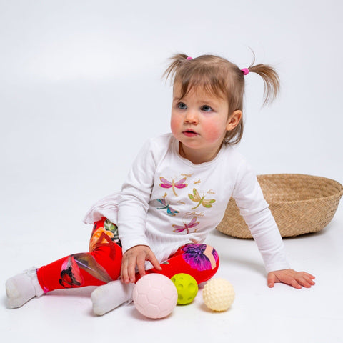 1. Baby sitting on a white background with pastel-colored sensory balls, wearing a white top with dragonfly print and red pants, enhancing sensory exploration.