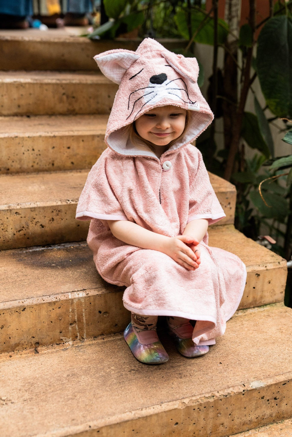 1. Toddler wearing RÄTT short-sleeve poncho CAT in pink, sitting on steps, showcasing hood with cat ears and face design