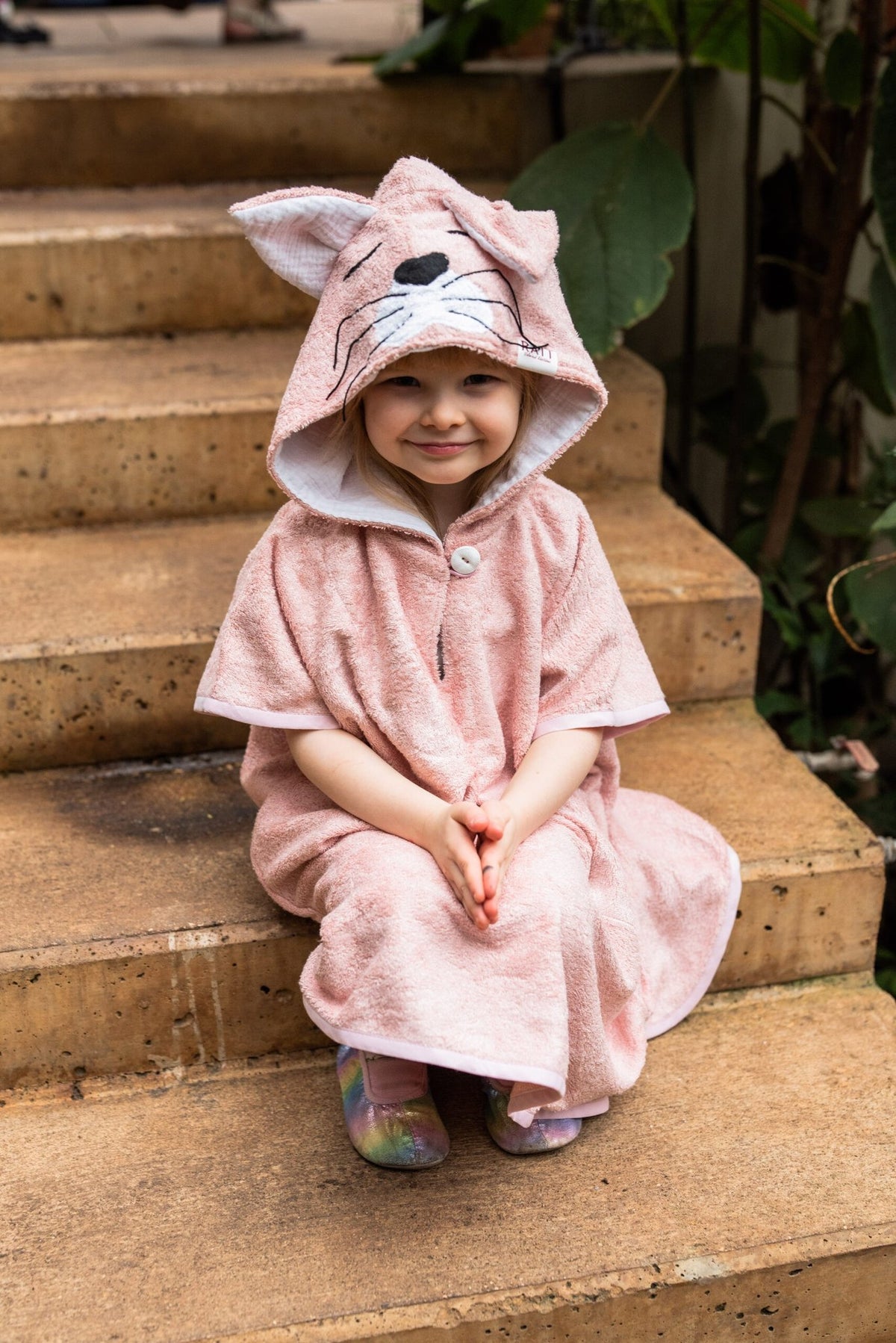 3. Toddler in RÄTT short-sleeve poncho CAT, sitting on steps, highlighting cozy design with hood and cat features