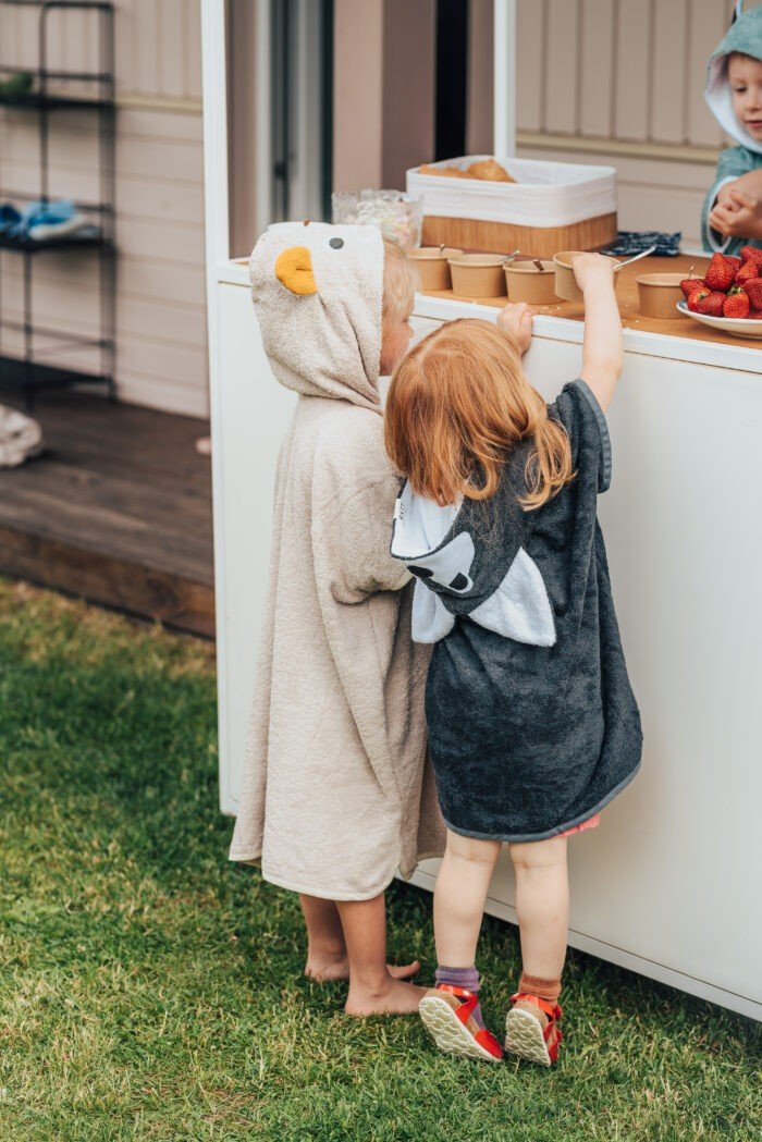 2. Two toddlers in RÄTT Short-Sleeve Poncho WOLF, one in black and one in beige, reaching for snacks outdoors