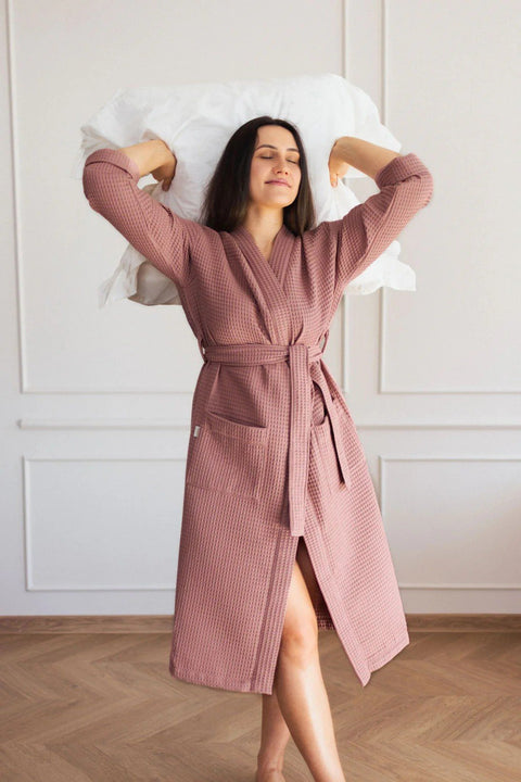 1. Woman wearing pink waffle fabric robe with belt and side pockets, holding pillow in bright room