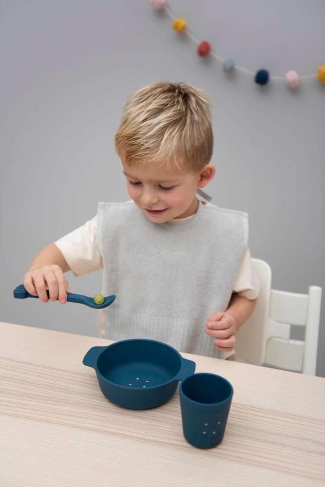 1. Young boy using blue silicone bowl and cup set at dining table, wearing a grey bib