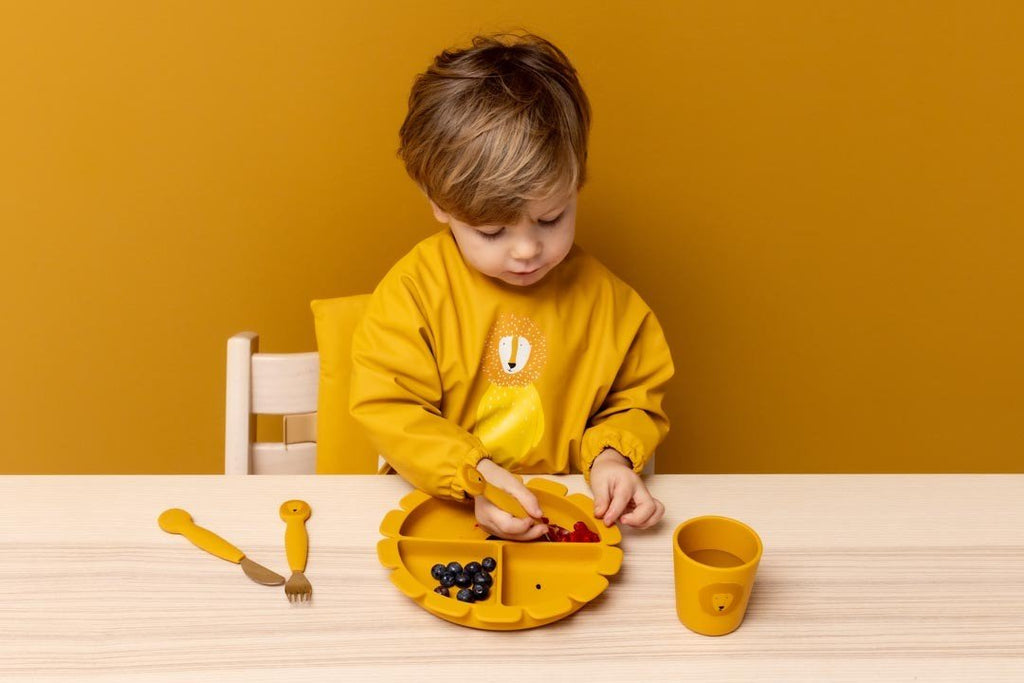 1. Child in yellow outfit using Trixie Baby silicone cup and plate set at wooden table