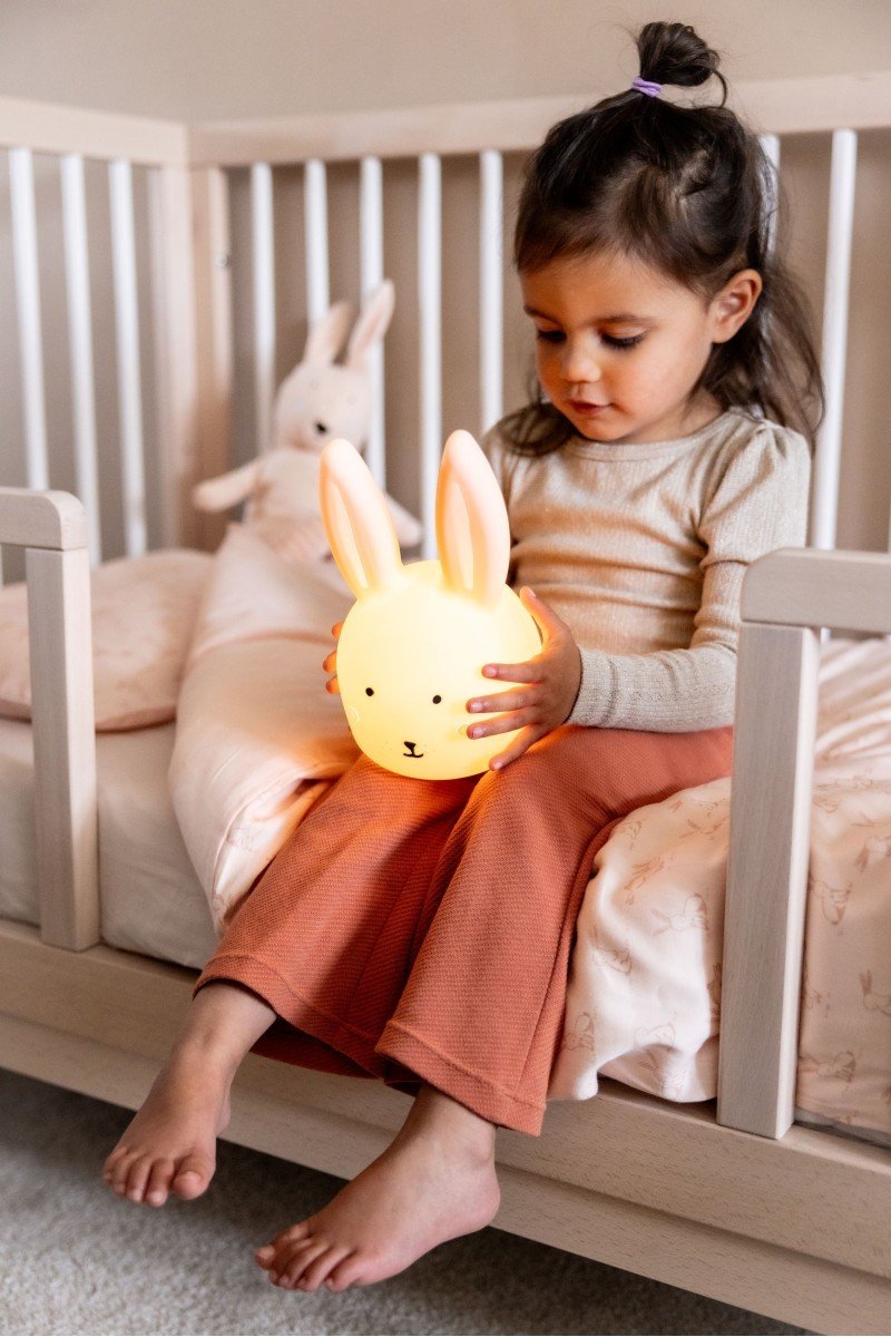 1. Young girl holding Trixie Baby Mrs. Rabbit silicone night light on bed in cozy bedroom setting
