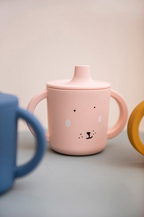 1. Pink silicone sippy cup with rabbit face design on a table with blue and yellow cups in the background