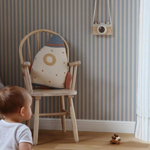 1. Child in a room with beige and blue striped wallpaper, wooden chair with rocket-shaped cushion, and hanging toy camera