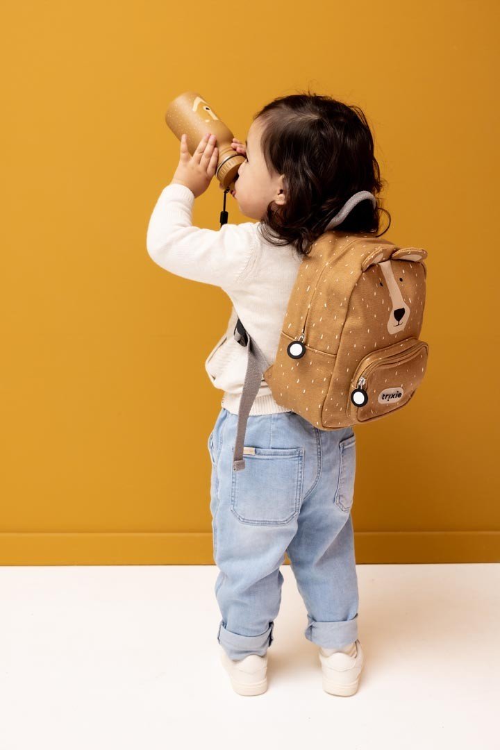 1. Child wearing Trixie Baby Mr. Bear small backpack, drinking from bottle, against mustard background
