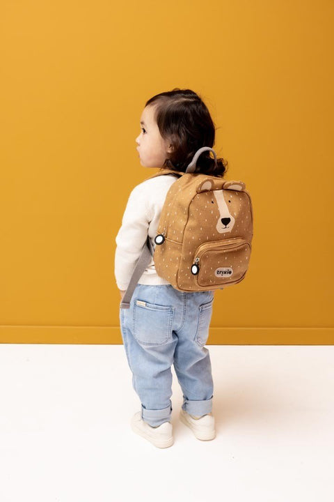 1. Child wearing Trixie Baby Mr. Bear small backpack, standing against mustard background