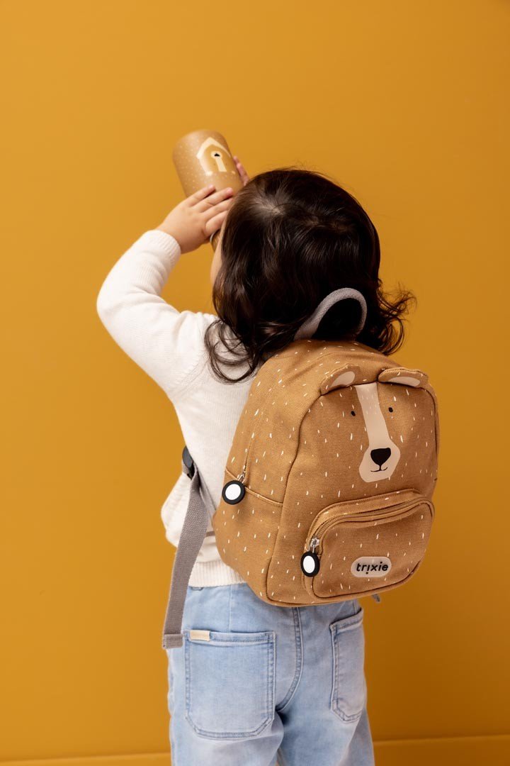 1. Child wearing Trixie Baby Mr. Bear small backpack, drinking from bottle, against mustard background